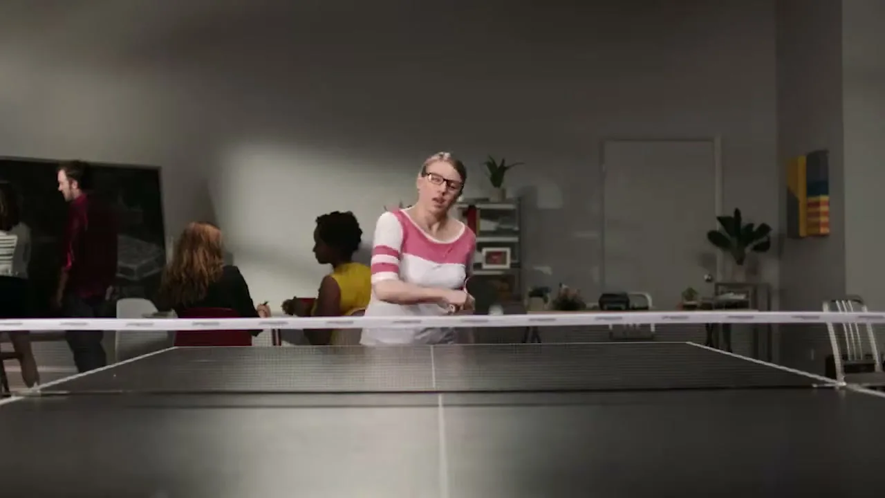A woman in glasses and a striped shirt plays ping pong in an office, with other people in the background.