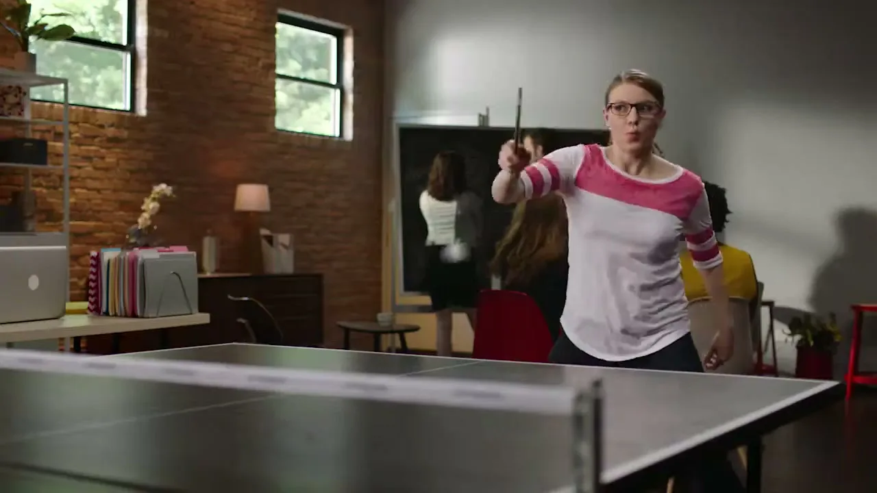 A woman in glasses and a pink and white shirt plays ping pong in an office, with other people in the background.