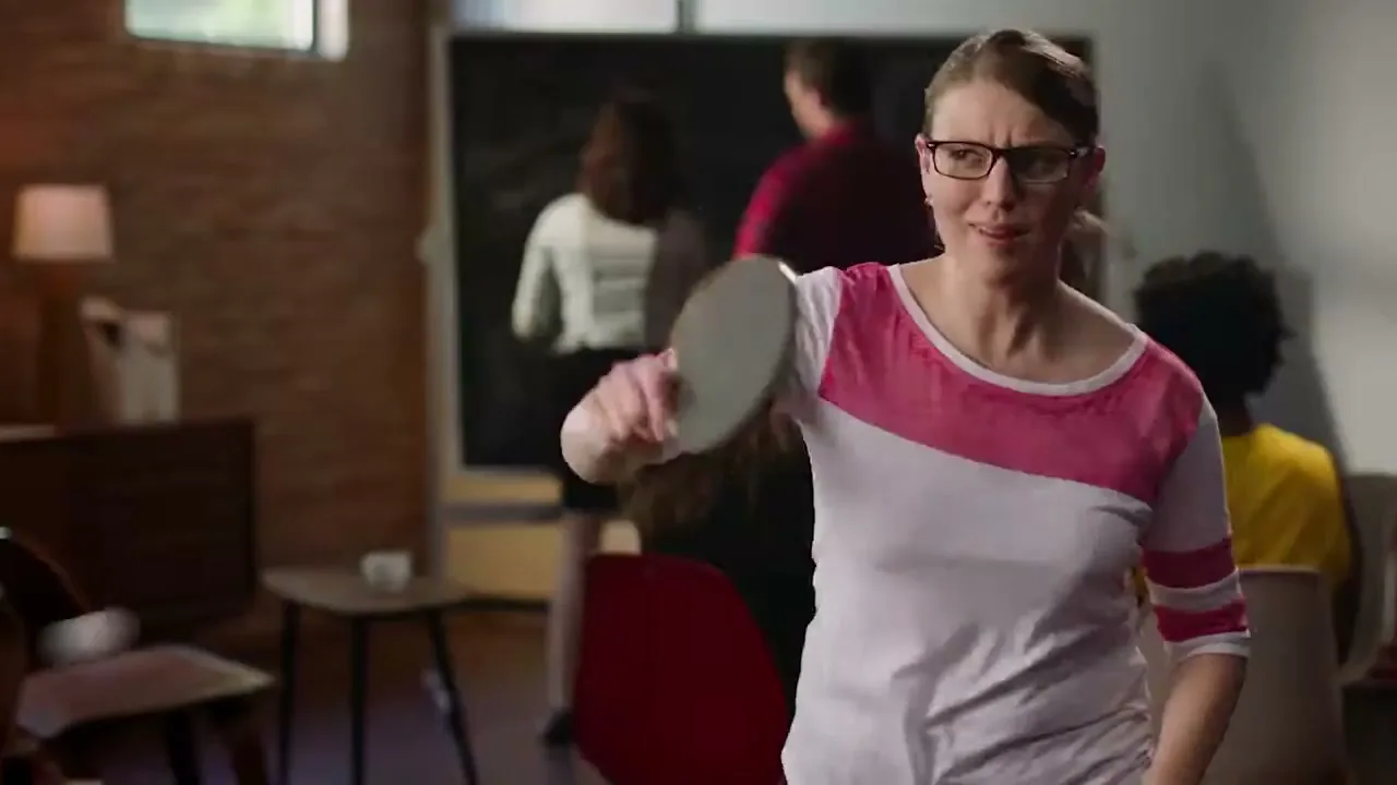 A woman in glasses and a pink and white shirt plays ping pong indoors, looking intently at the ball.
