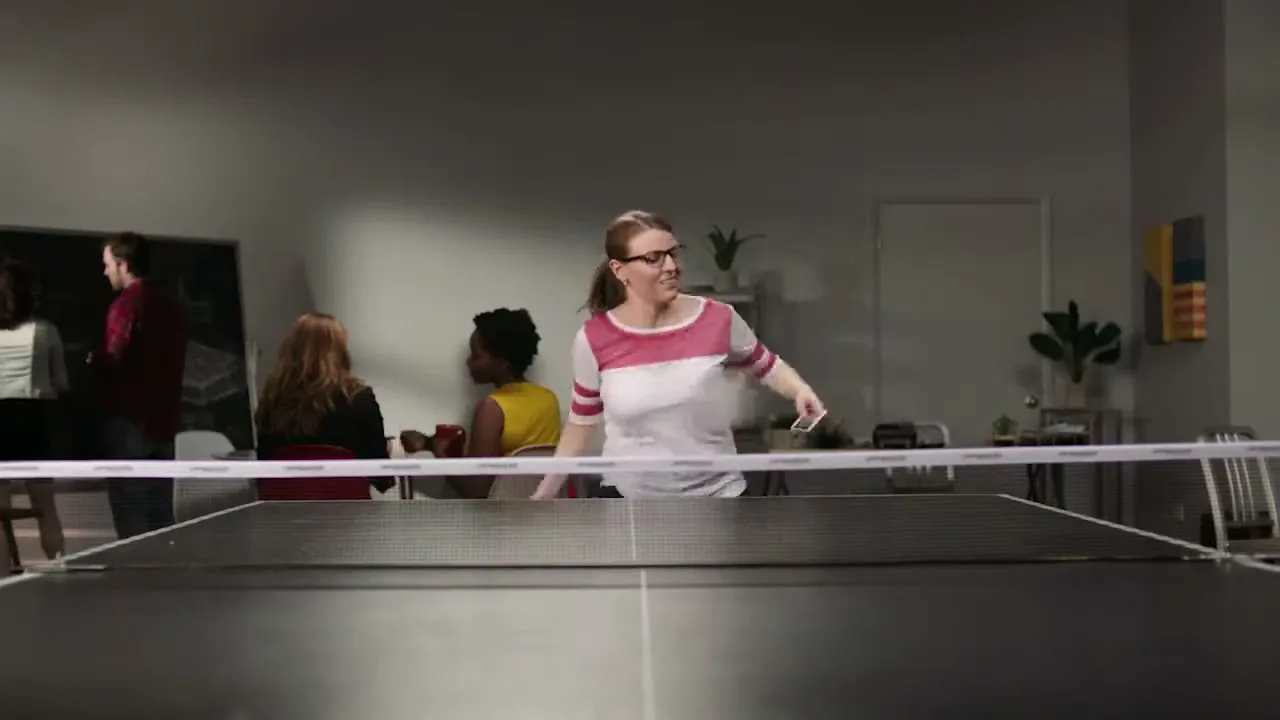 A woman in glasses and a striped shirt plays ping pong in an office, holding a phone and paddle, while others are in the background.
