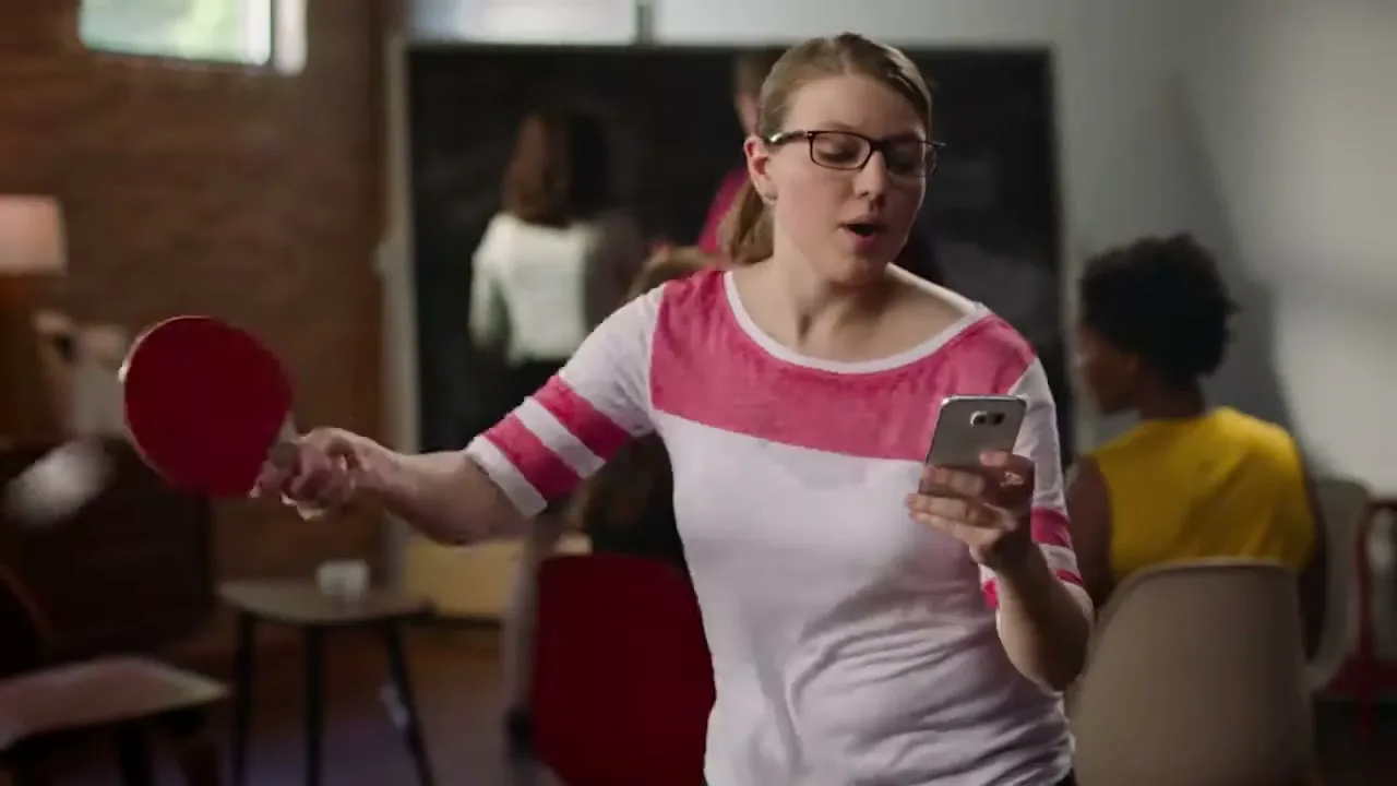 A young woman in glasses and a pink and white shirt plays ping pong while looking at her phone in an office.