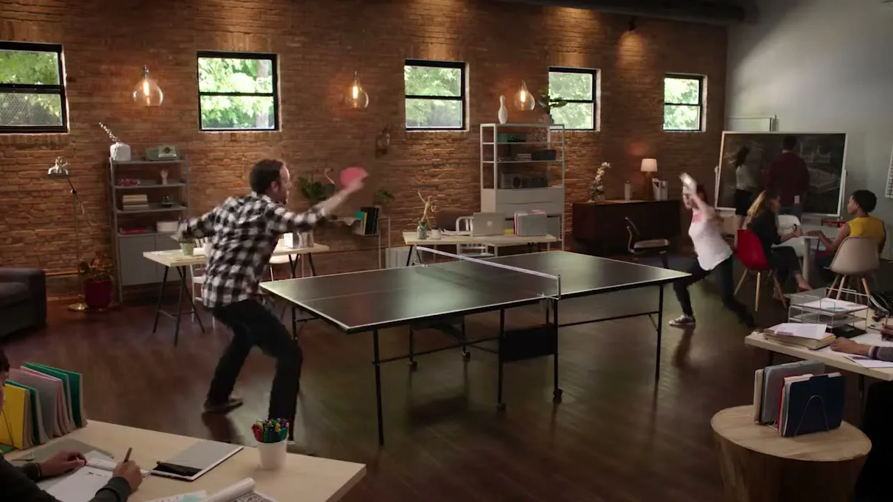 A man and a woman play ping pong in a modern office with brick walls and large windows, while other colleagues work or watch in the background.