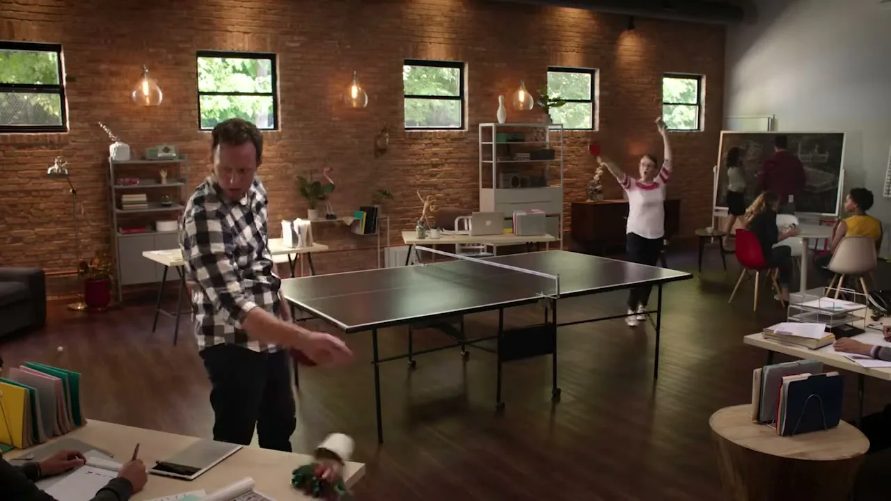 A man in a plaid shirt and a woman in a white shirt play ping pong in a modern office with exposed brick walls and large windows.