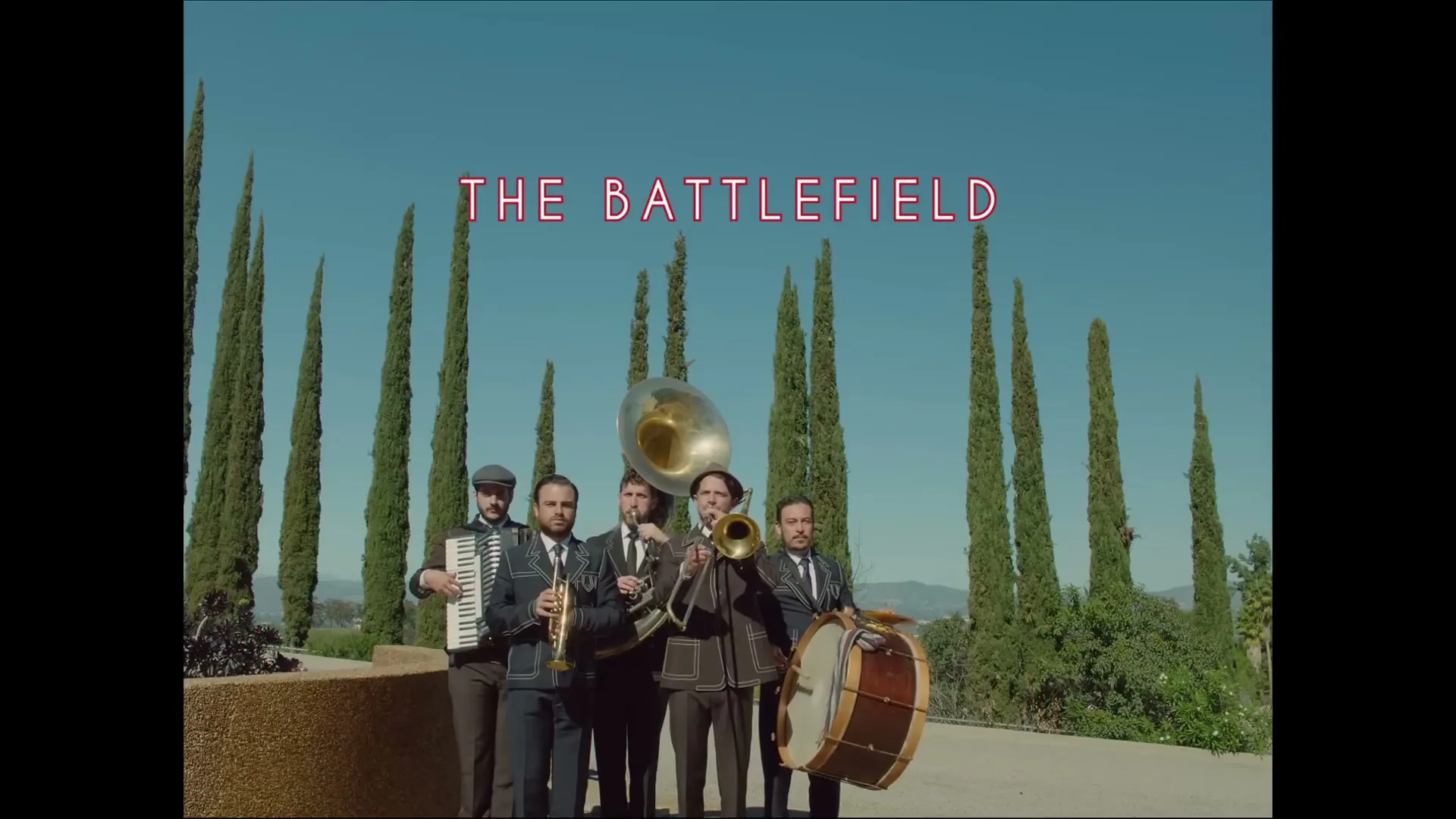 A band of five men in suits plays instruments in front of tall, slender trees under a clear blue sky, with "THE BATTLEFIELD" text overlay.