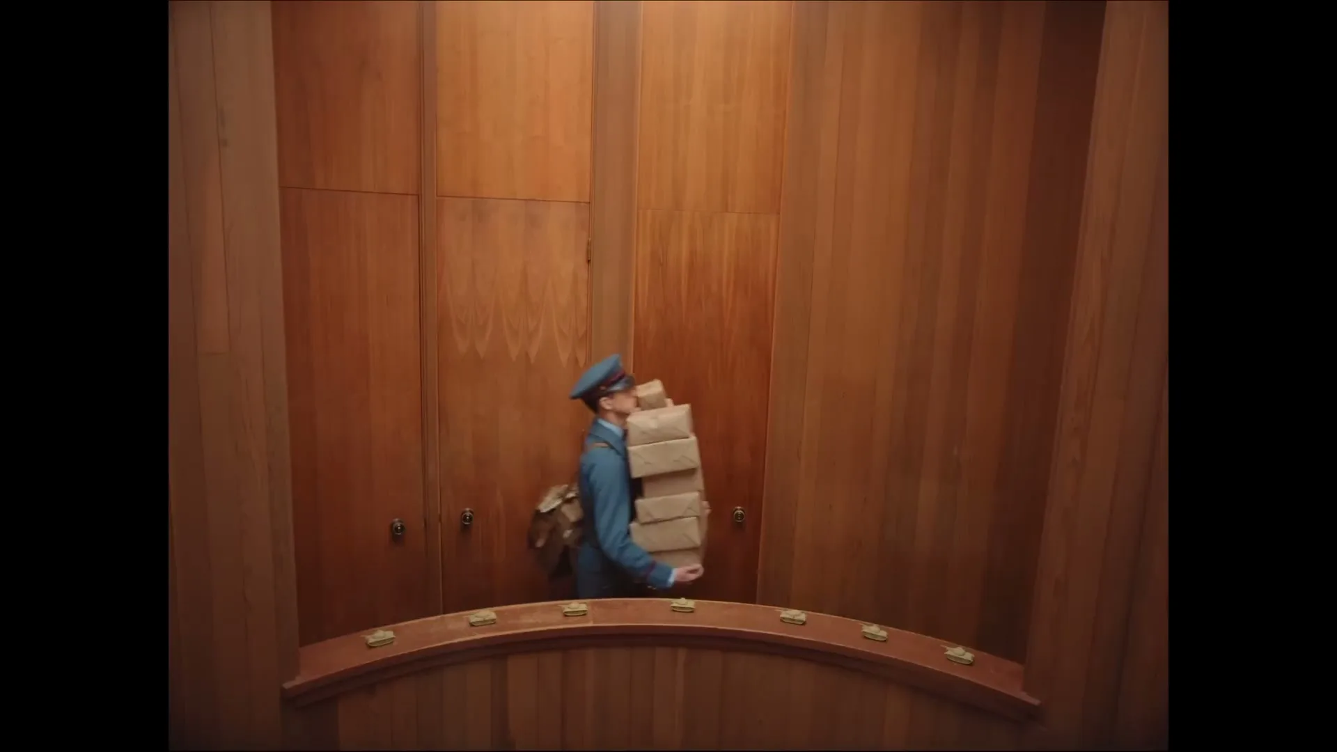 A male postman in a blue uniform struggles to carry a tall stack of packages through a circular, wood-paneled room.