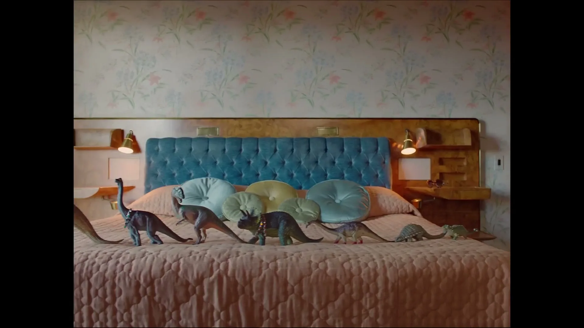 A line of toy dinosaurs, some with jewelry, walk across a pink quilted bed with a blue tufted headboard in a bedroom with floral wallpaper.