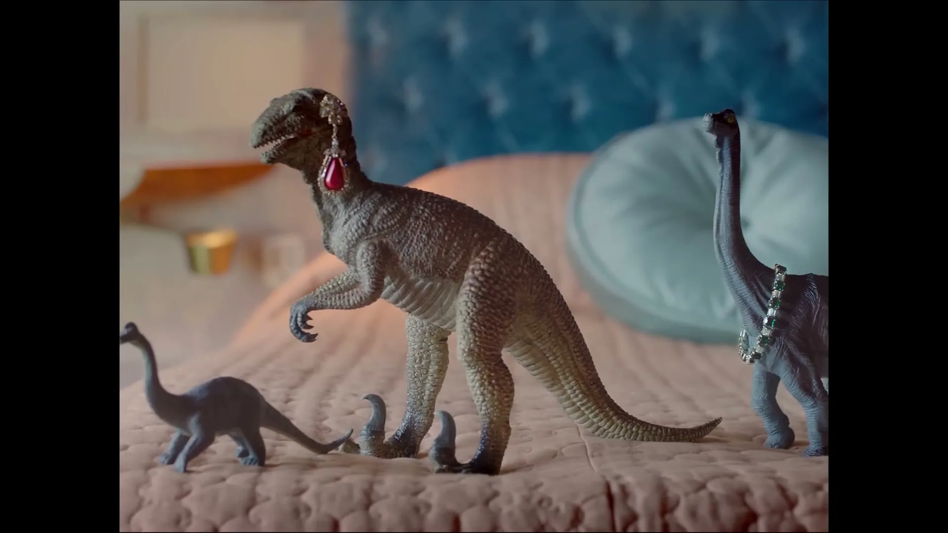 Three toy dinosaurs, one wearing a red earring and another a green necklace, stand on a pink bedspread in a bedroom.
