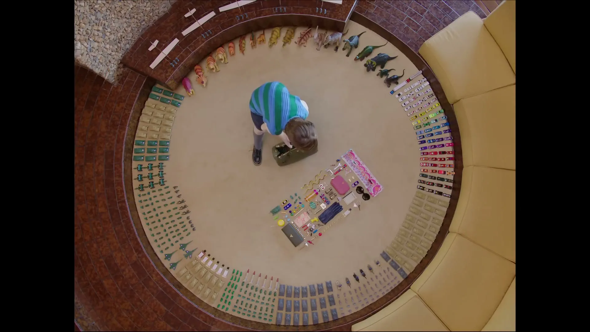 A child in a striped shirt plays with a collection of toys, including dinosaurs and small accessories, arranged in a circular pattern on a carpeted floor.