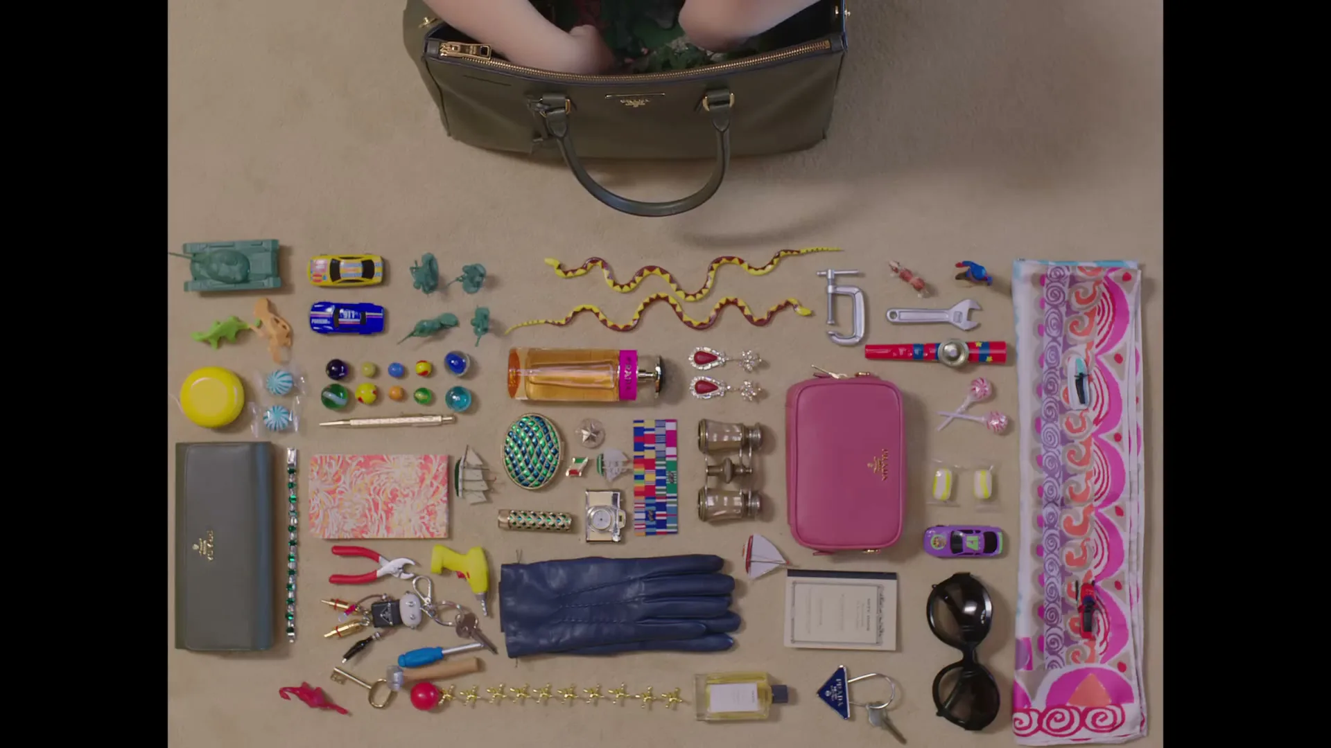 A woman's hands are seen from above, placing a toy soldier into a Prada handbag, surrounded by an array of personal items laid out on a beige surface.
