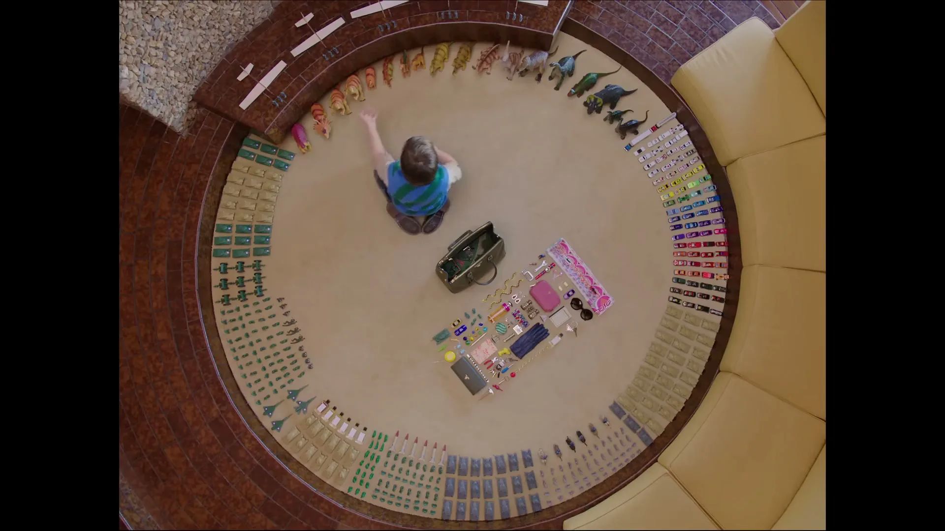 A young boy sits on a circular rug, surrounded by toys and accessories, as he reaches for something in a high-angle overhead shot.