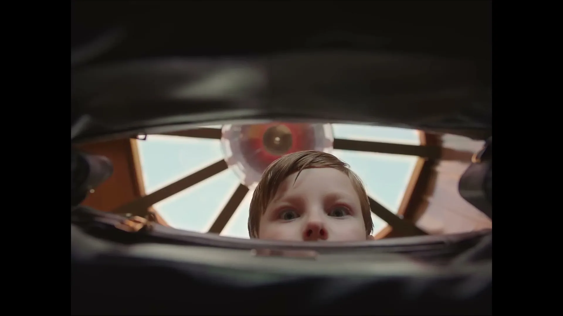 A young boy with light brown hair looks up into the camera, which is positioned inside a dark bag, with a colorful toy visible behind him.