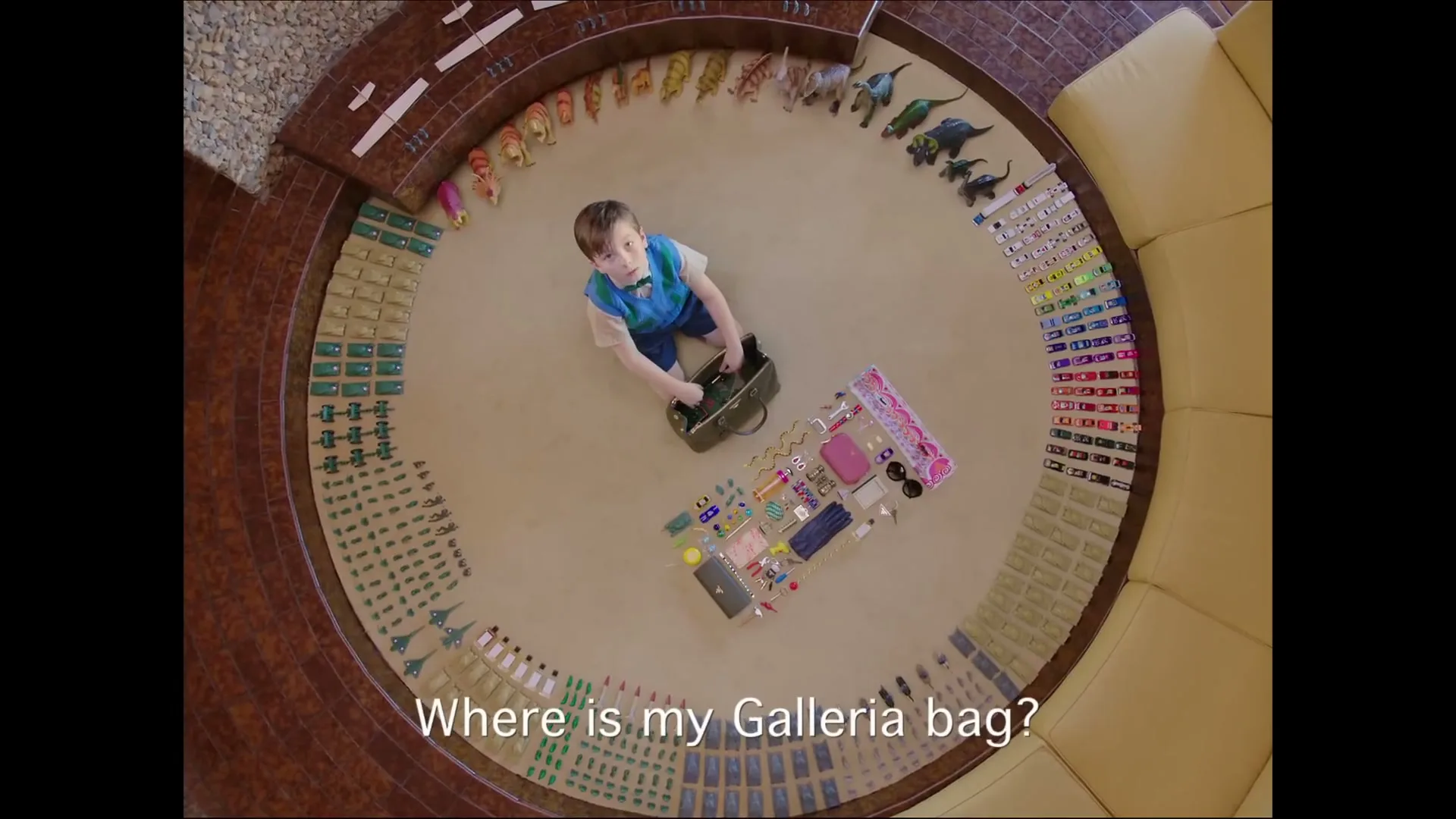 A young boy in a blue vest and bow tie looks up at the camera, surrounded by a circular arrangement of toys and accessories on a carpeted floor.