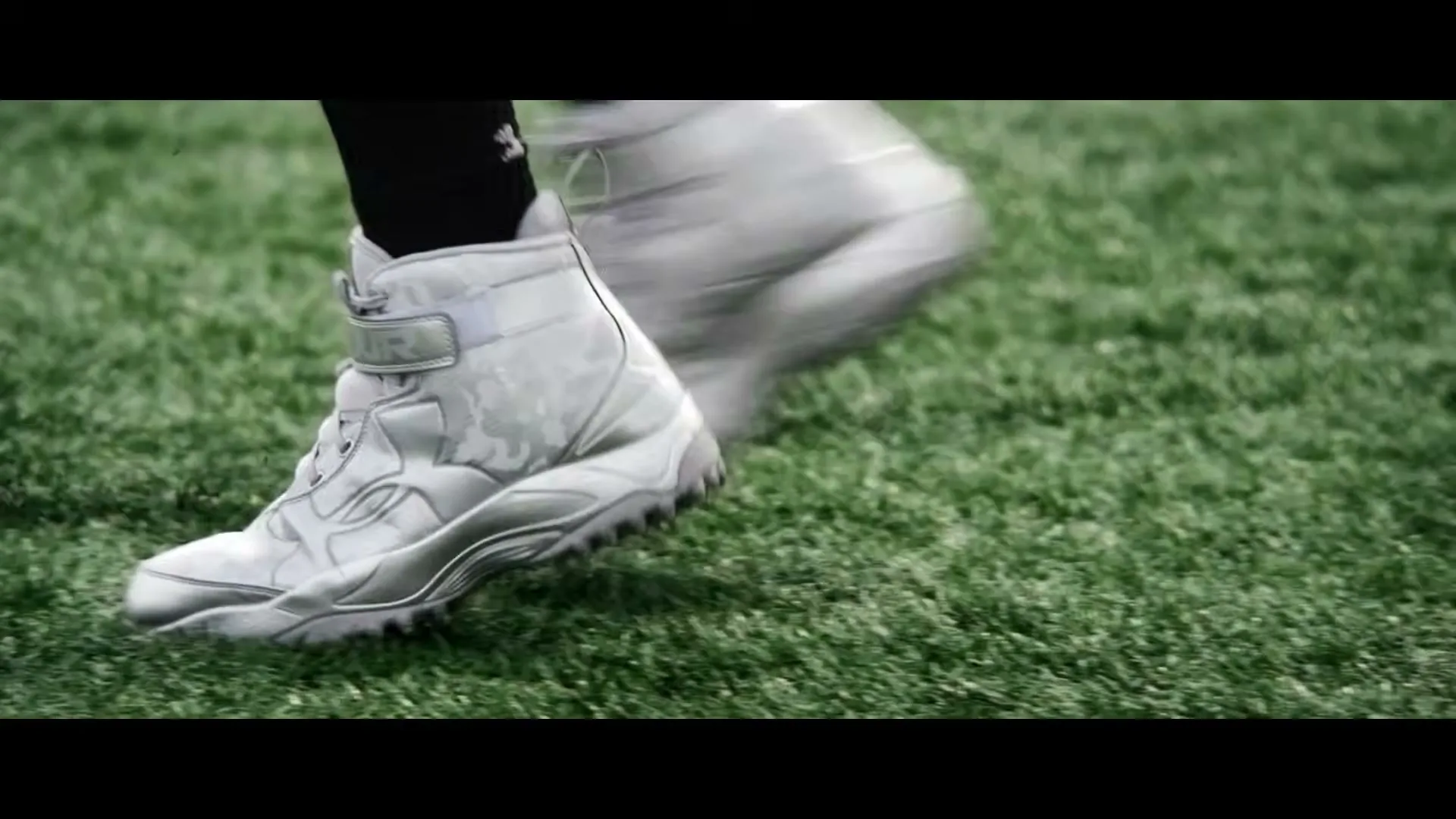A person's feet in white cleats run across a green football field, kicking up small bits of turf.