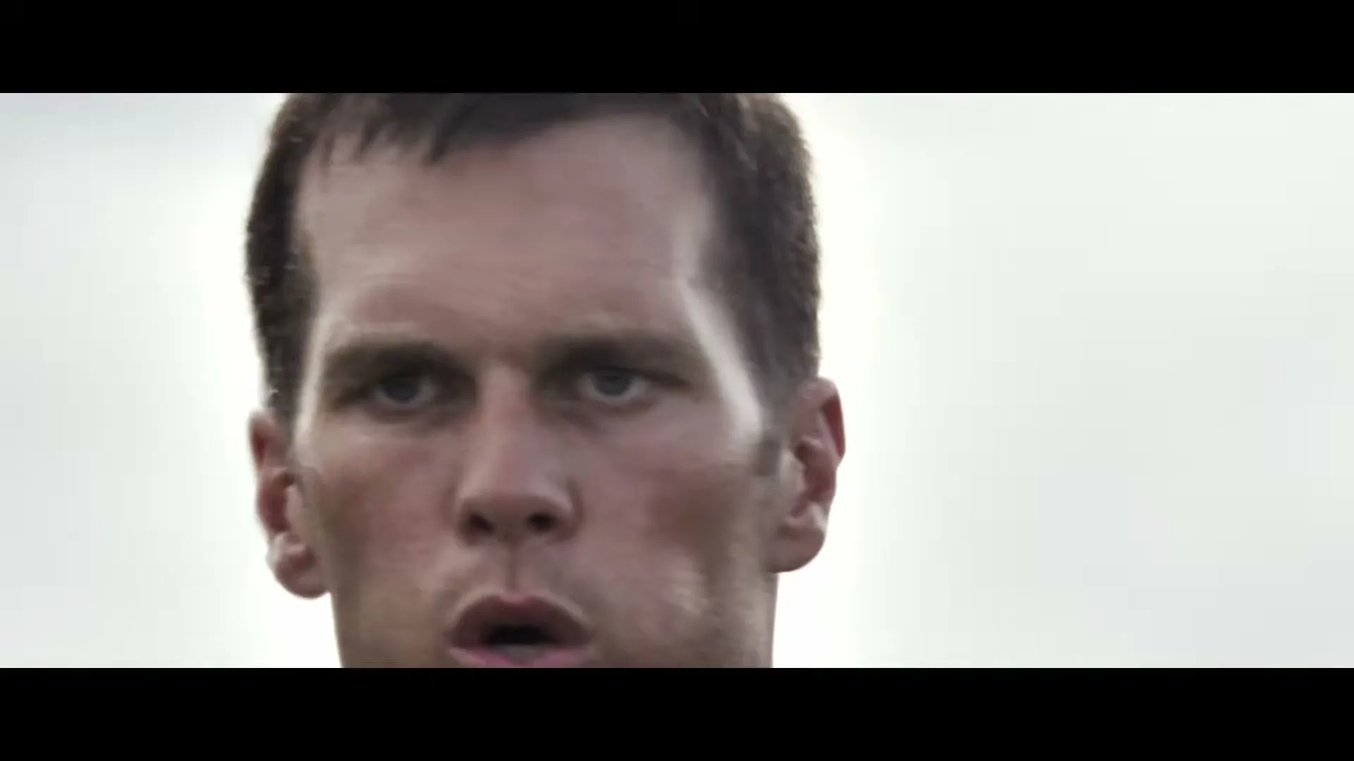 A football player, Tom Brady, wearing a black shirt, looks intensely forward on a field during the day.