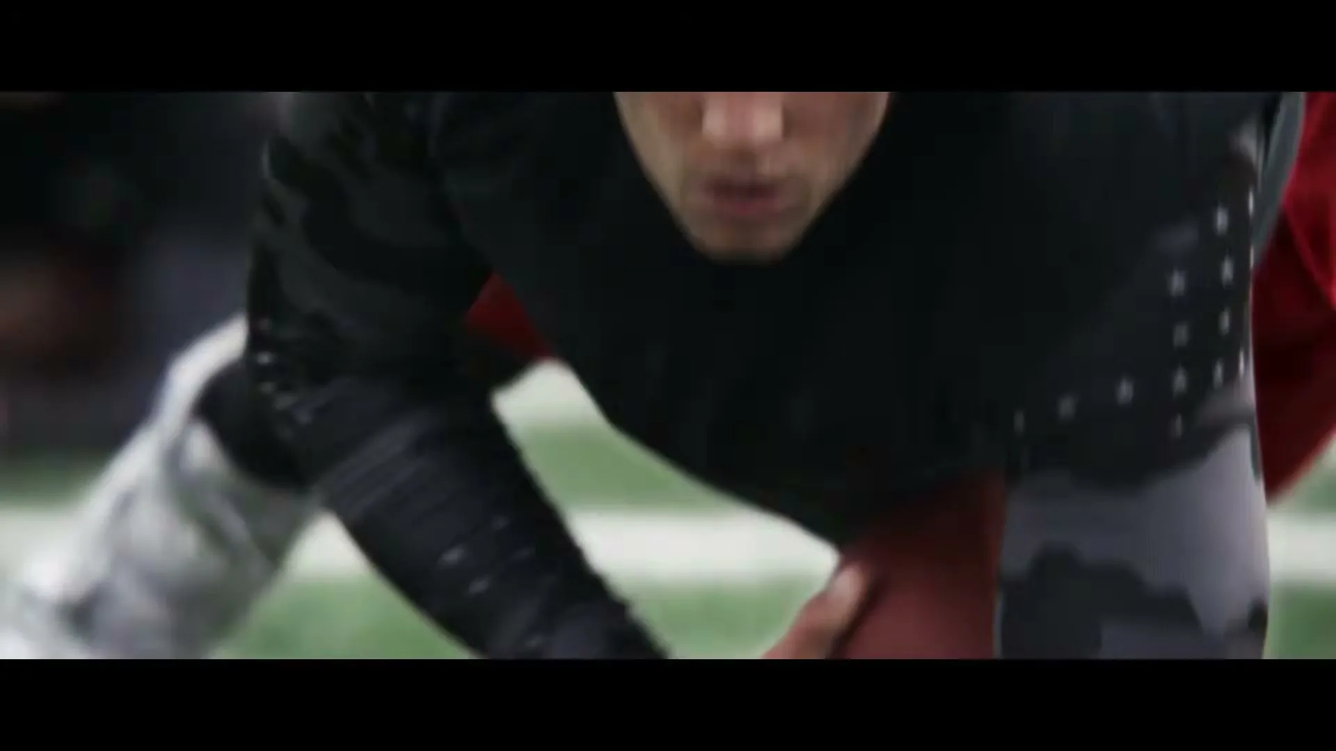A male athlete in a black shirt and white glove with a logo is shown in a medium closeup, focused on the ground, holding a football on a green field.
