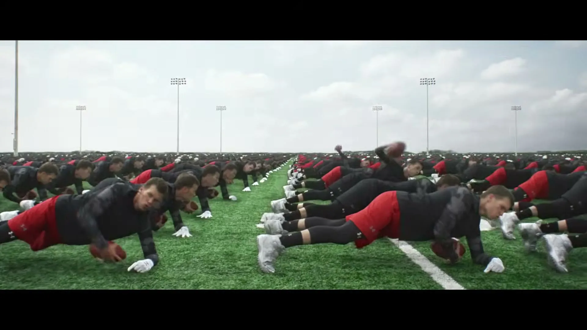 A wide shot shows hundreds of identical men, all resembling Tom Brady, doing push-ups on a vast green football field under a cloudy sky.
