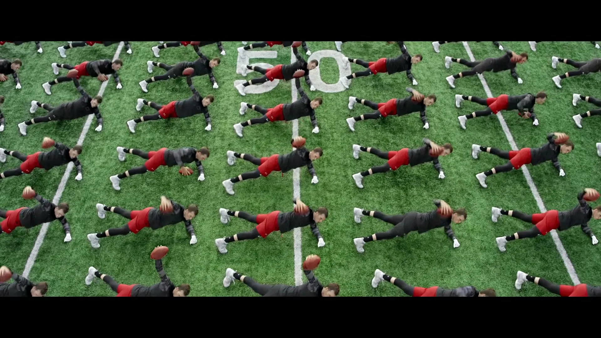 A wide shot shows many clones of a male athlete in black and red sportswear doing push-ups on a green football field.
