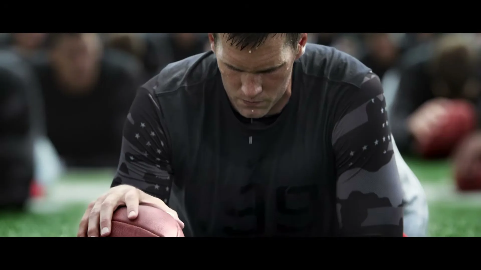 Tom Brady, a male athlete, is seen in a medium shot, sweating and focused, with his hand on a football on a green field, surrounded by other athletes.
