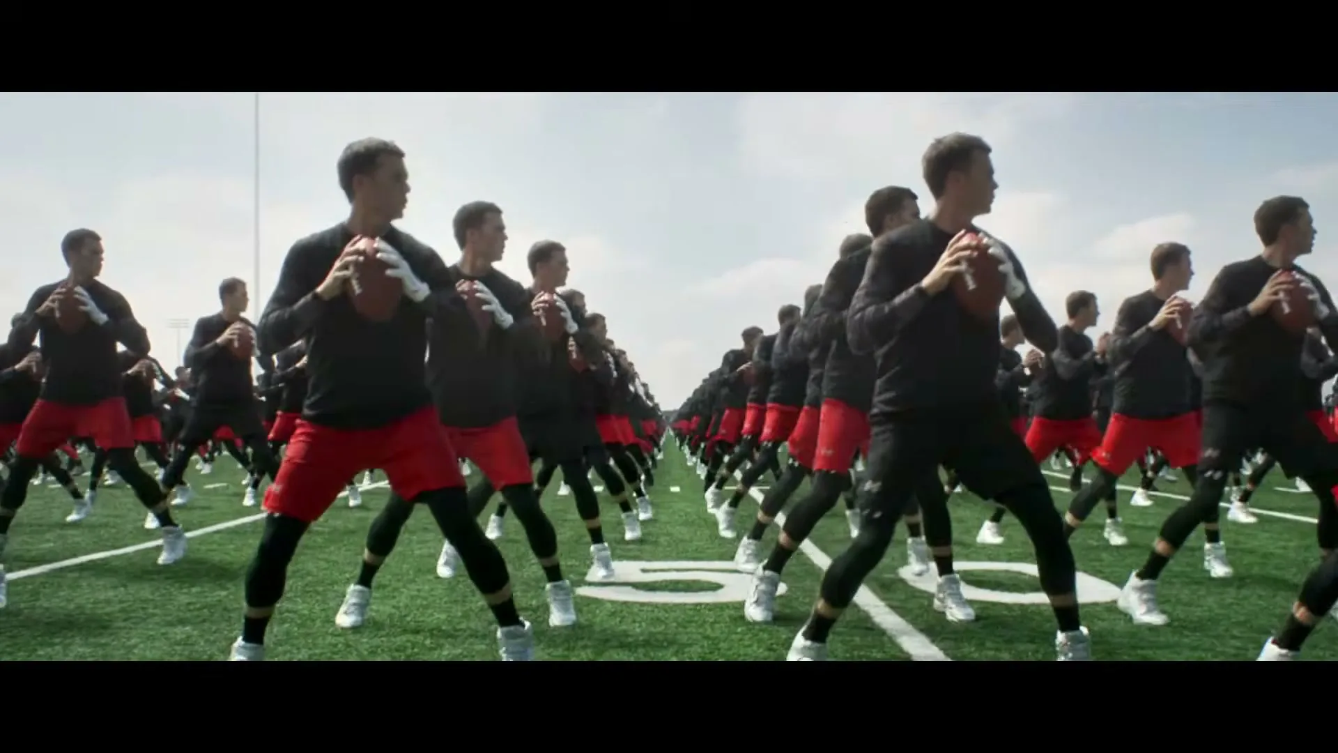 A wide shot shows many clones of Tom Brady in black shirts and red shorts, holding footballs on a green football field under a bright sky.