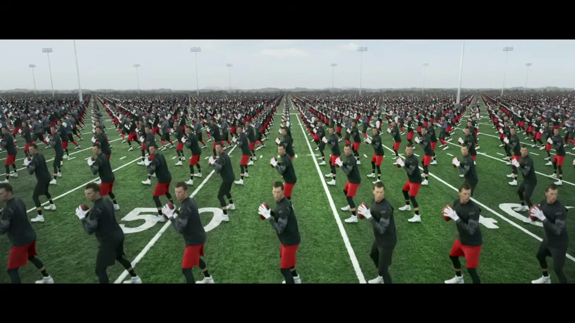 A wide shot shows hundreds of Tom Brady clones in black shirts and red shorts, holding footballs and performing drills on an endless football field.