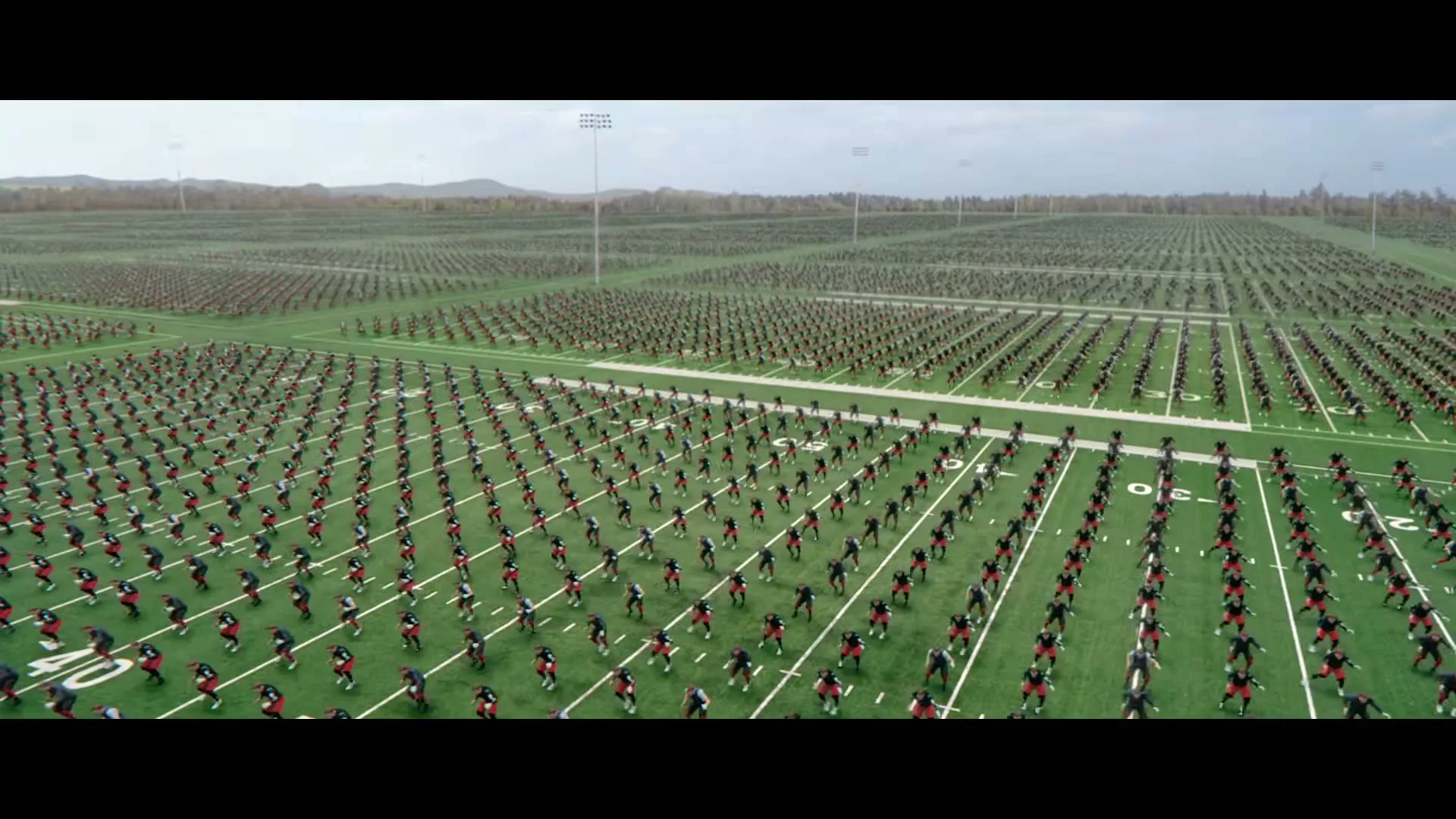 A wide shot of a football field filled with hundreds of football players in black and red uniforms, all in a ready stance, then a closeup of a player's feet and hands on the grass.