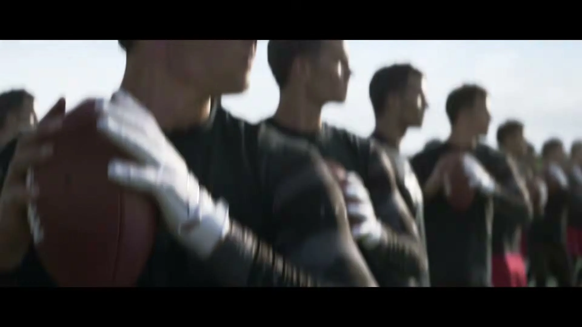 Tom Brady and other football players in black shirts and red shorts stand in a line on a sunny field, holding footballs.