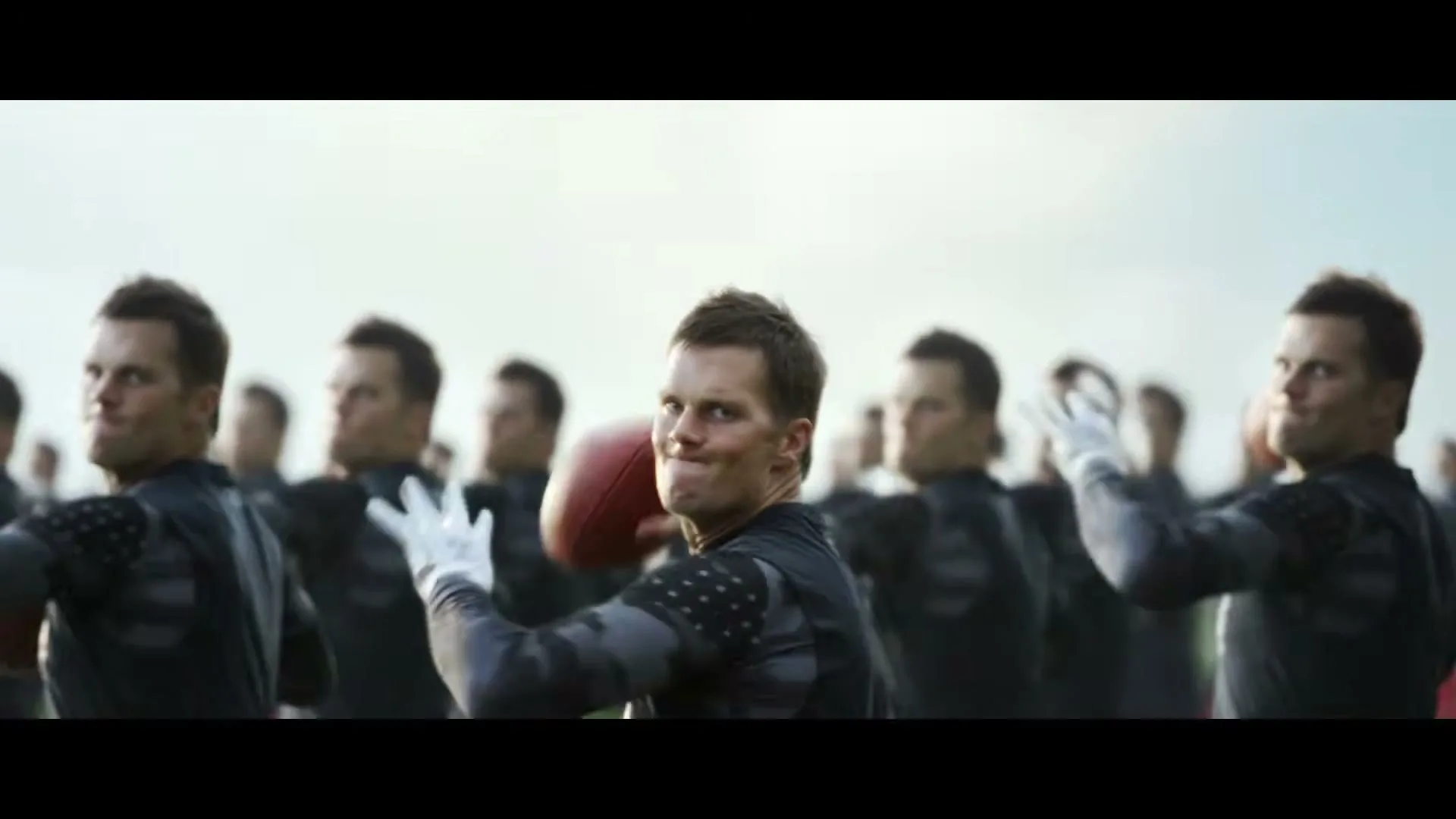 Tom Brady, wearing a black and grey athletic shirt, throws a football while surrounded by many clones of himself on a field.
