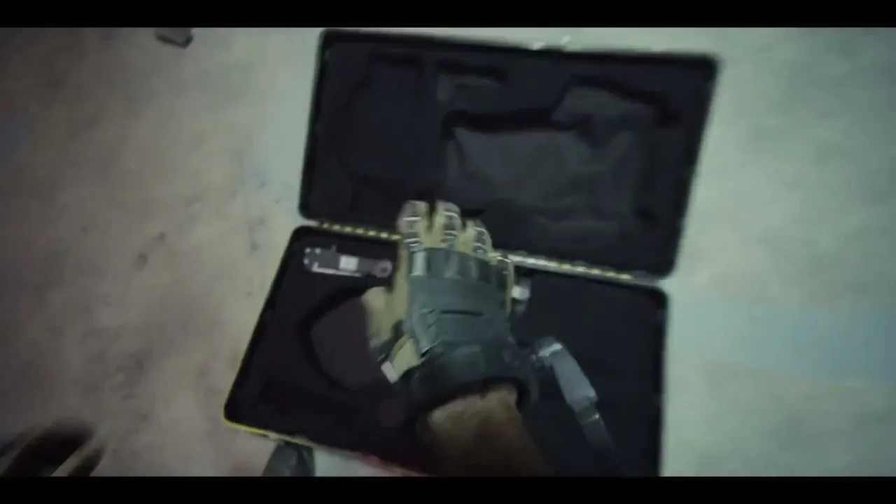 A man's hands assemble a futuristic, weaponized gauntlet from pieces stored in a black case on a concrete floor.