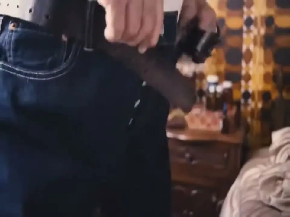 A person's hands are seen buttoning up a pair of dark blue jeans and putting on a brown leather belt in a bedroom.