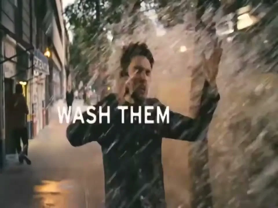 A man in a dark jacket talks on a phone while walking on a city sidewalk, then gets splashed by water from a passing bus.