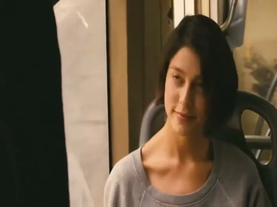 A young woman with short dark hair, wearing a grey sweatshirt, sits and looks to her left in an interior setting.