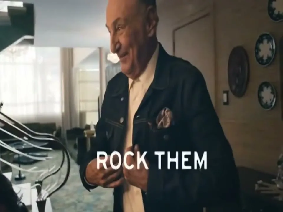 A smiling older man in a denim jacket adjusts his shirt in a modern living room, with "ROCK THEM" text on screen.