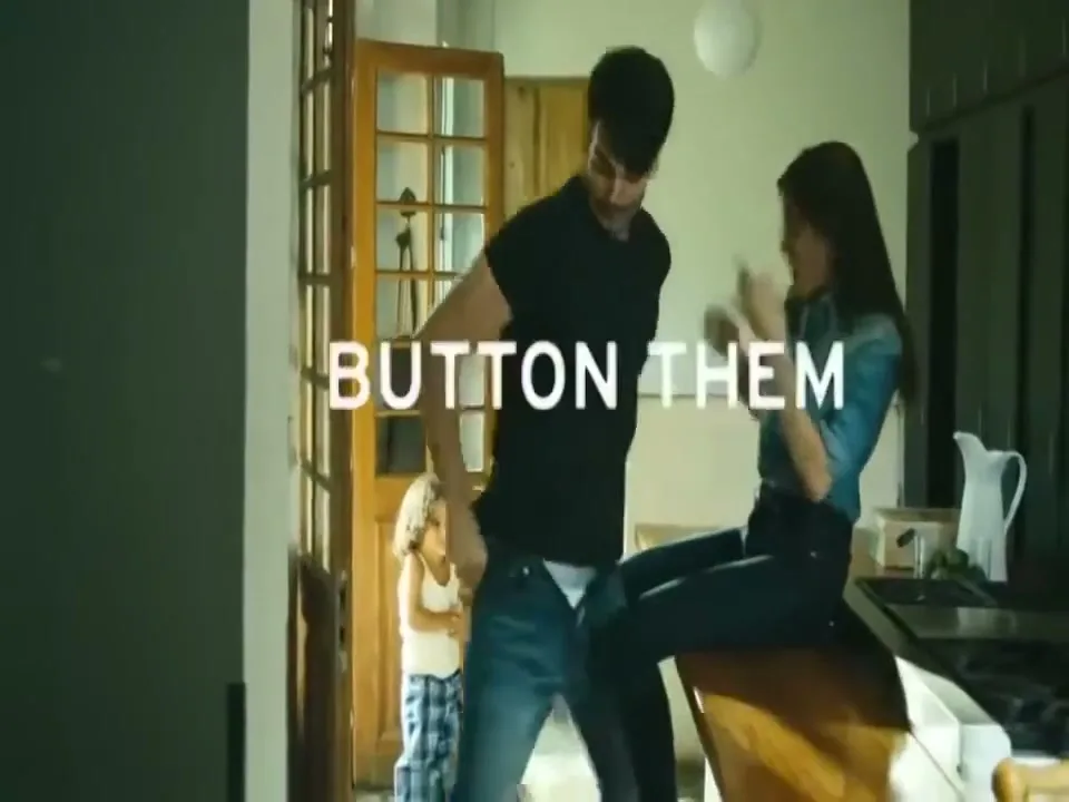 A man and woman kiss in a kitchen while a child watches, then the man buttons his jeans as text appears on screen.