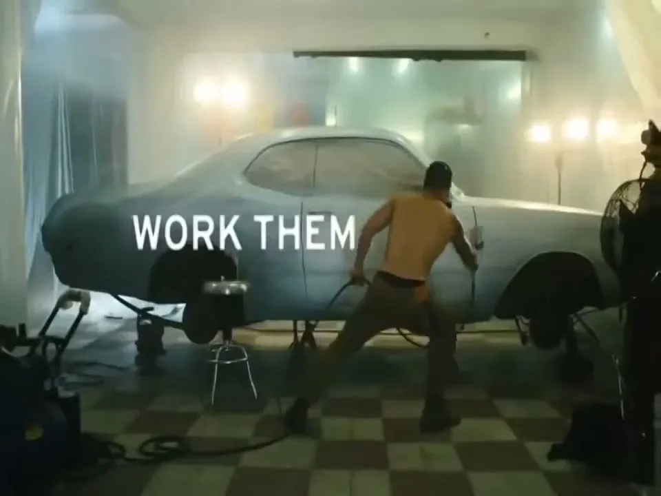 A shirtless man in pants spray paints a car in a garage, with the words "WORK THEM" projected onto the car.