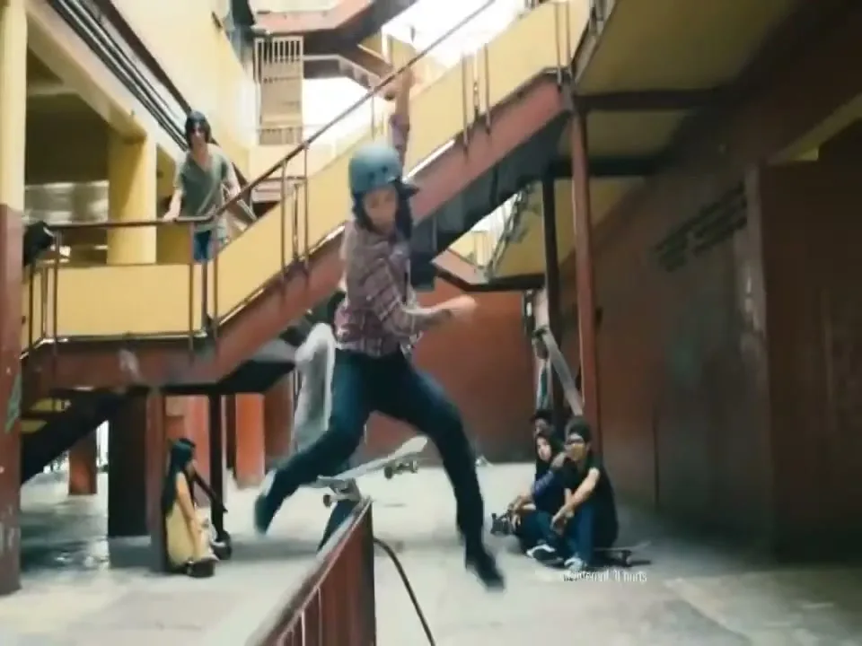 A young male skateboarder in a helmet jumps over a rail, while other young people watch in an industrial-looking building.