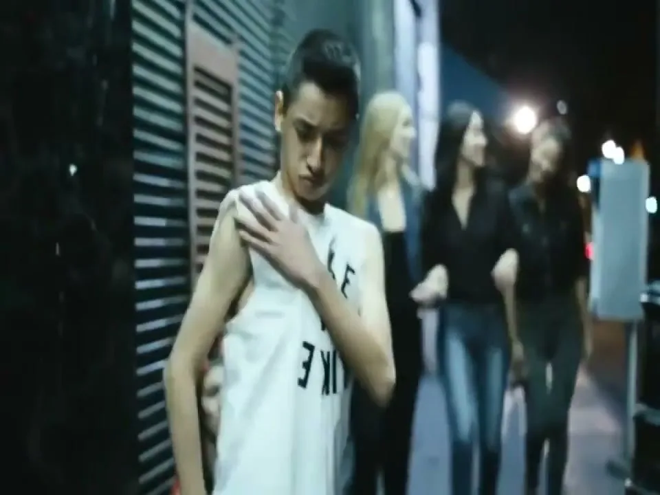 A young male in a white tank top dances awkwardly on a sidewalk at night, while three women walk past him in the background.