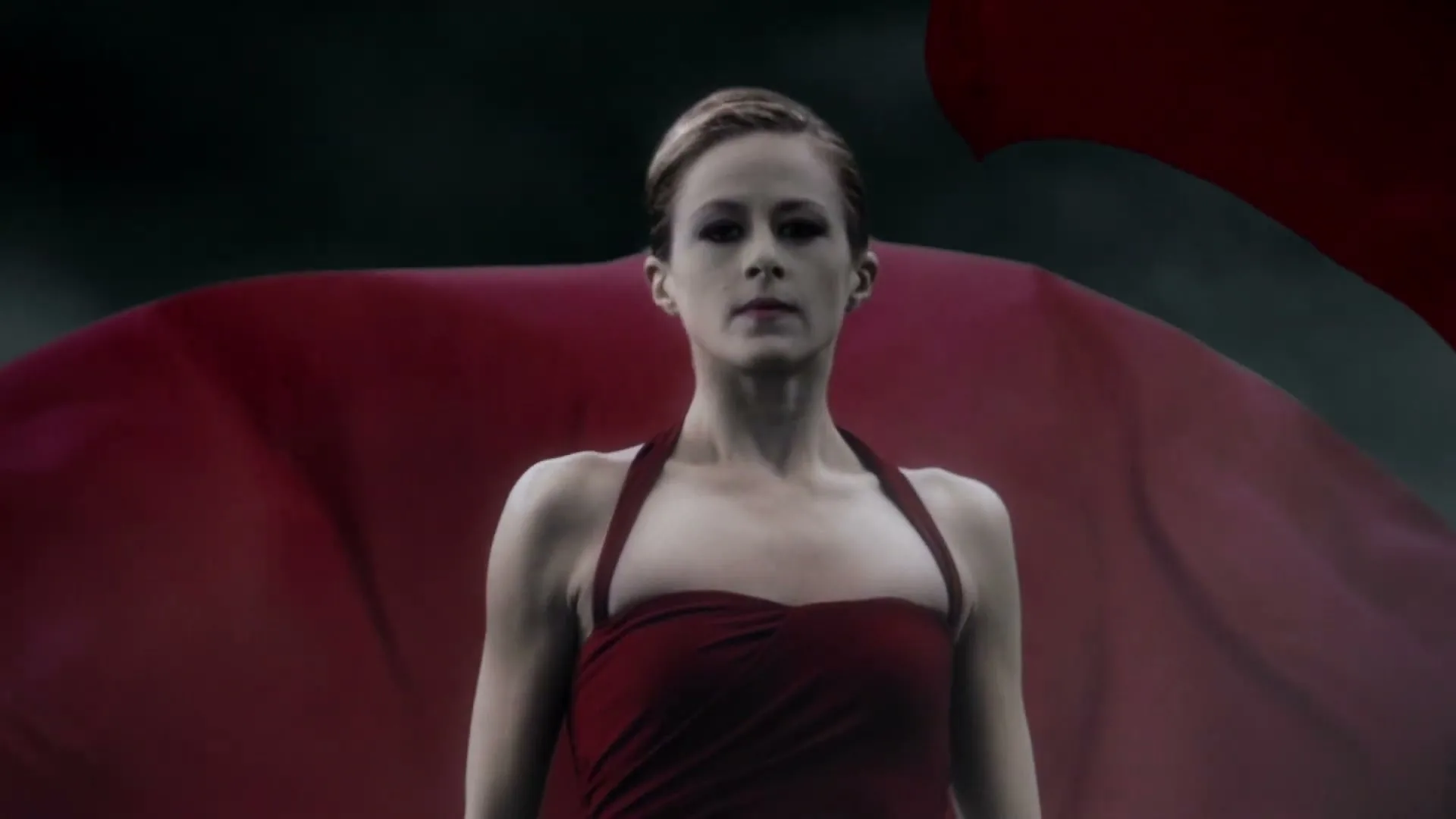 A female ballet dancer with short hair and a red dress stands still as a large red fabric billows around her in a dark, moody setting.