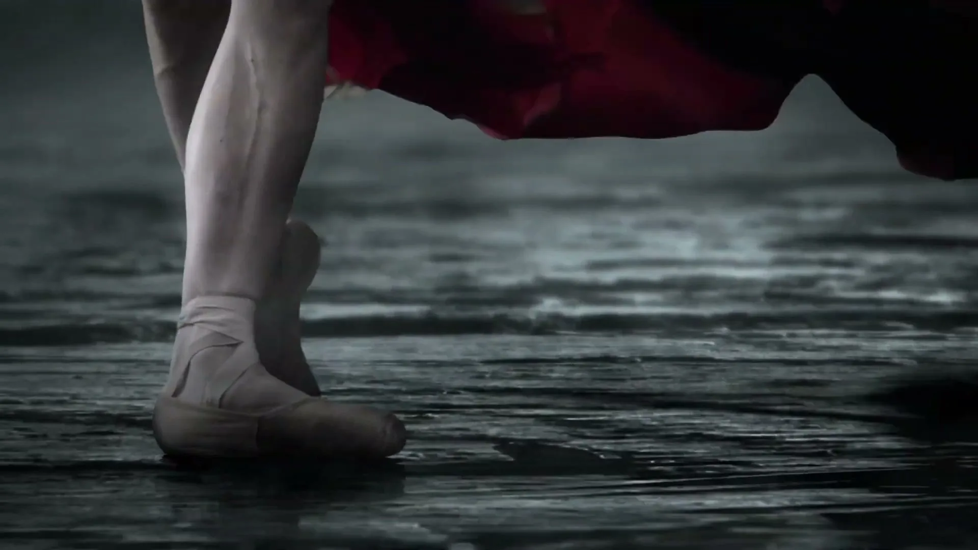 A ballerina's feet in pointe shoes, wearing a red dress, dance gracefully on a dark, wet surface.