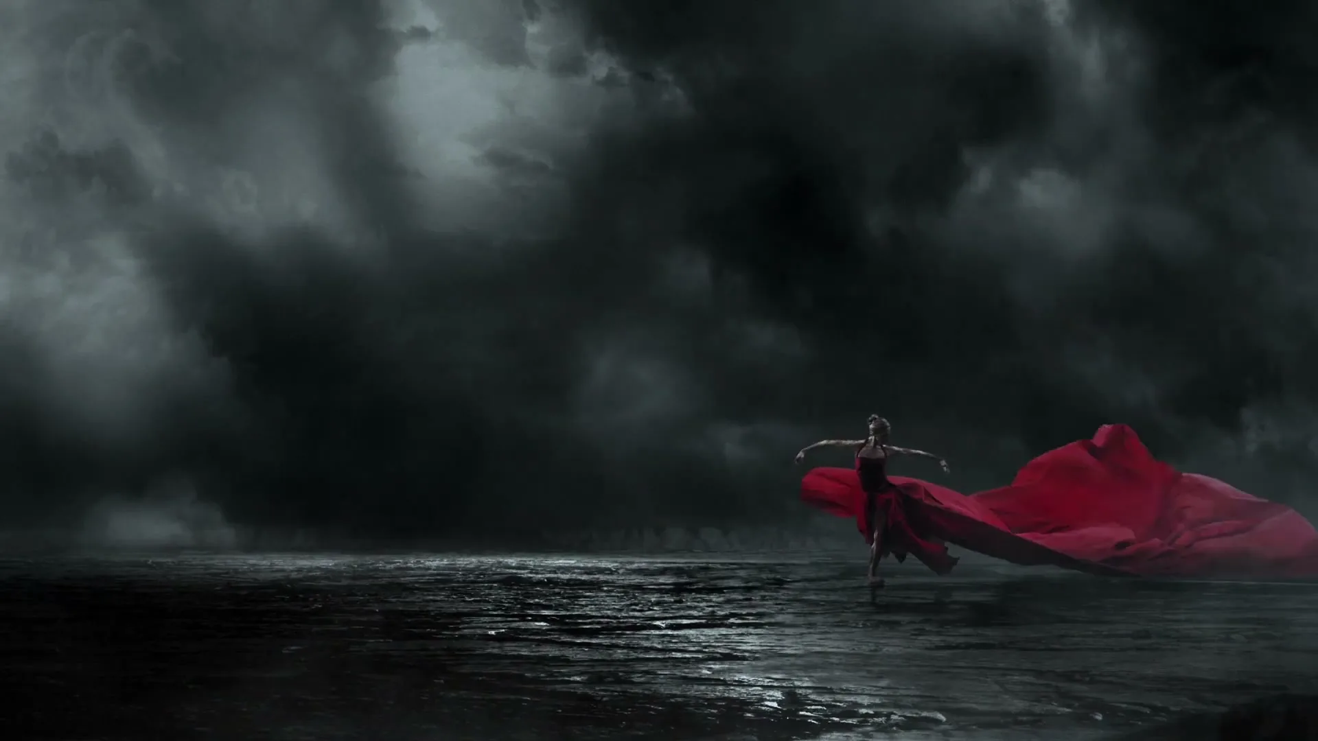 A female ballet dancer in a black leotard and a long red flowing skirt dances on a dark, wet surface under a stormy sky.