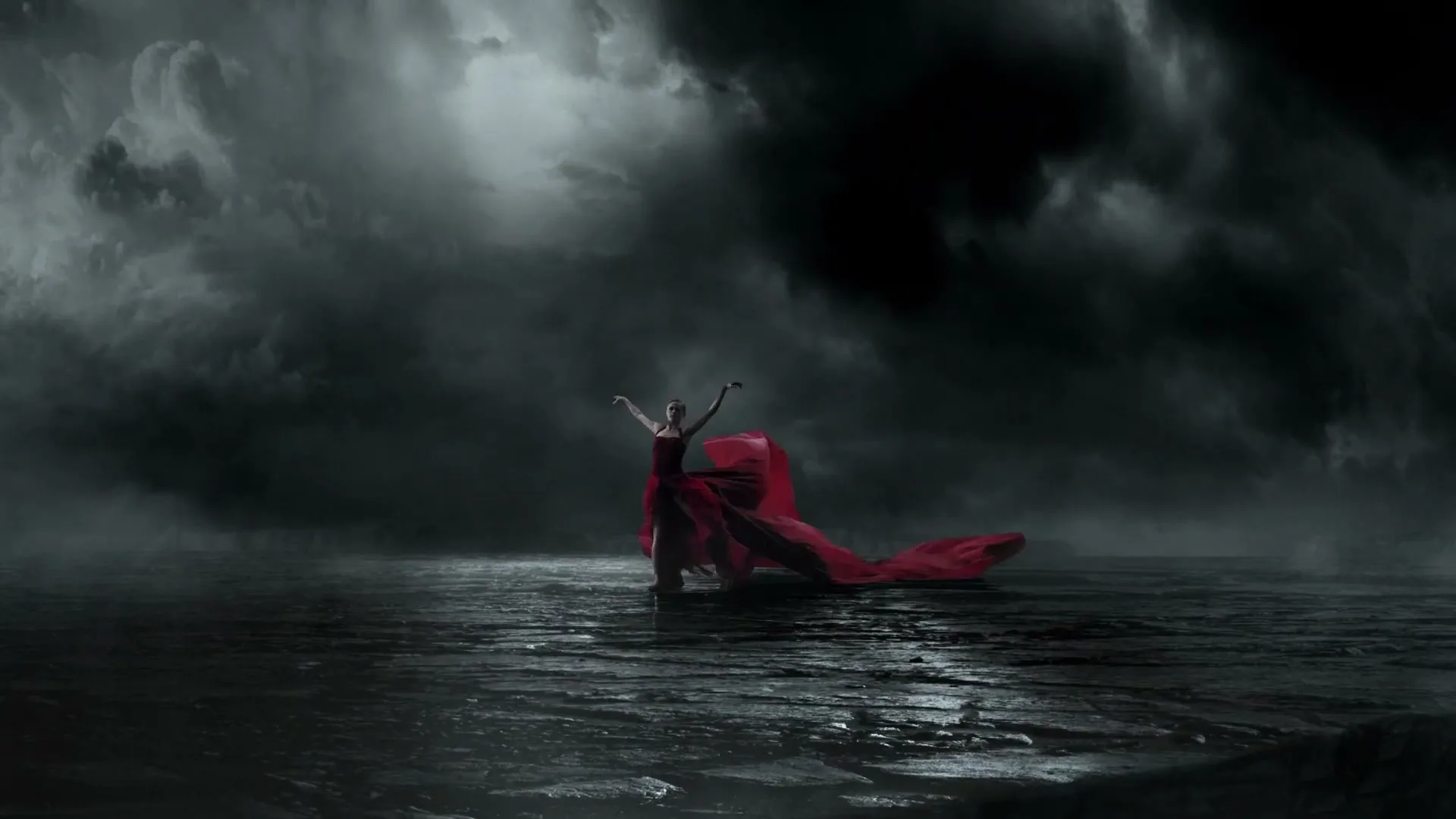 A female ballet dancer in a flowing red dress performs on a dark, reflective surface under a stormy, cloudy sky.