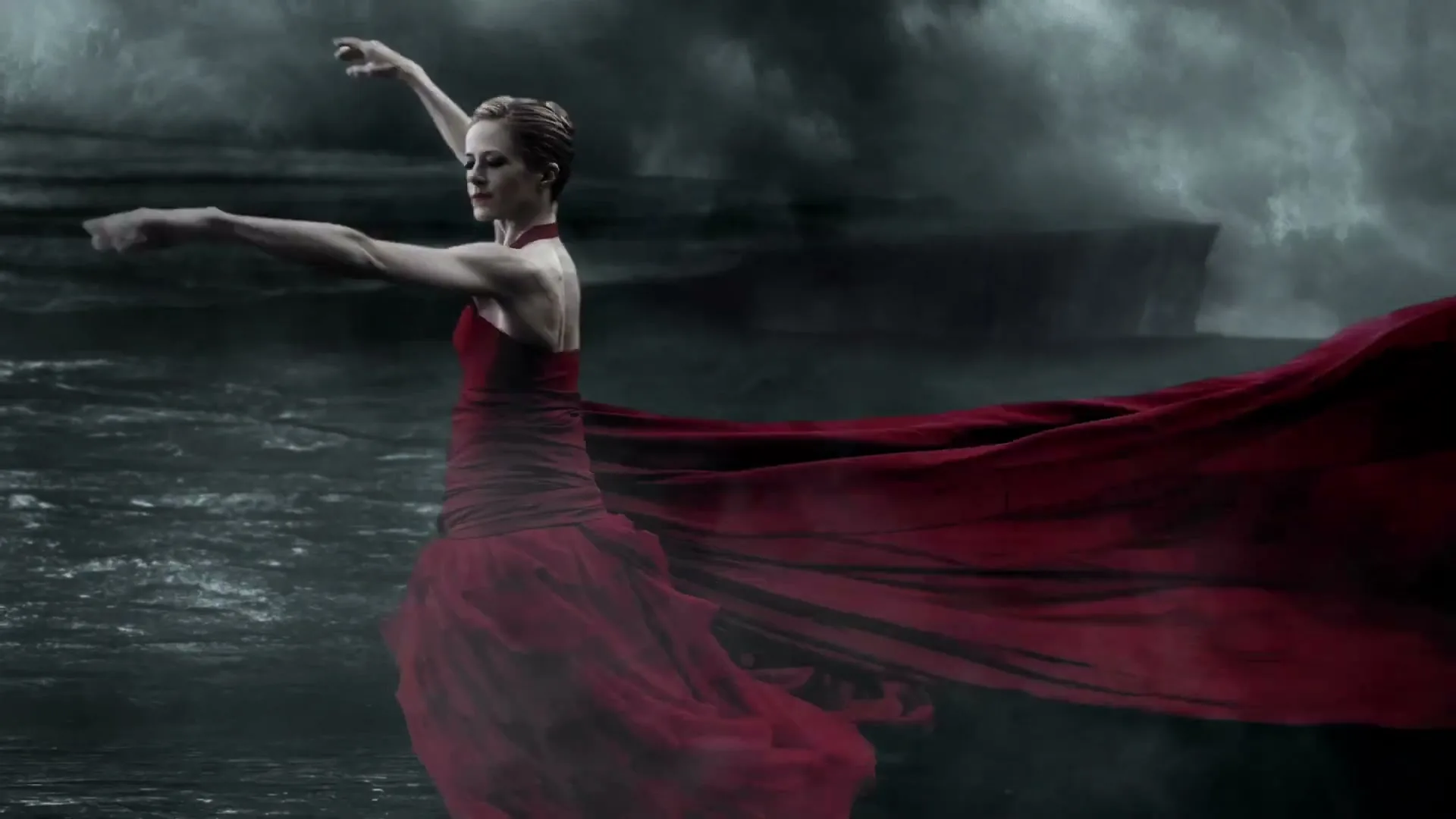 A female ballet dancer in a flowing red dress performs a graceful movement against a dramatic, stormy, dark water background.