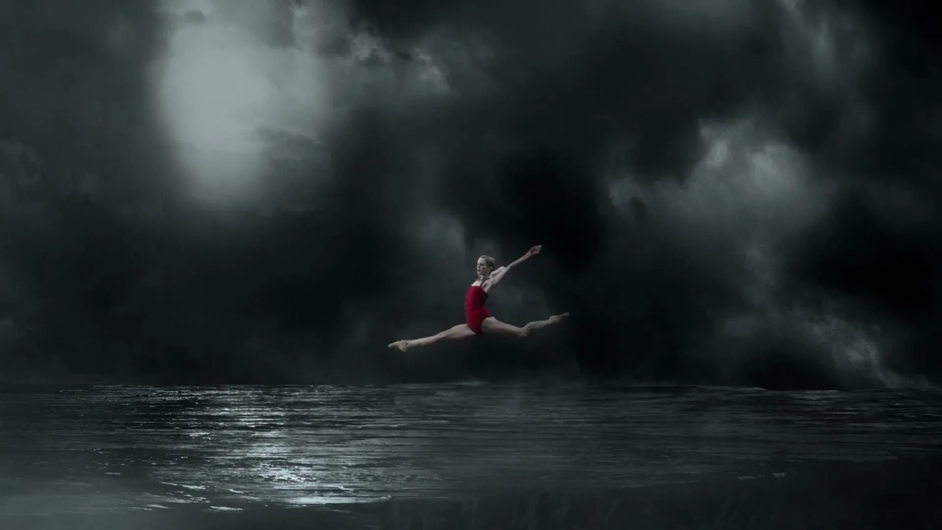 A female ballet dancer in a red leotard performs a grand jeté on a reflective surface, surrounded by dark, stormy clouds.
