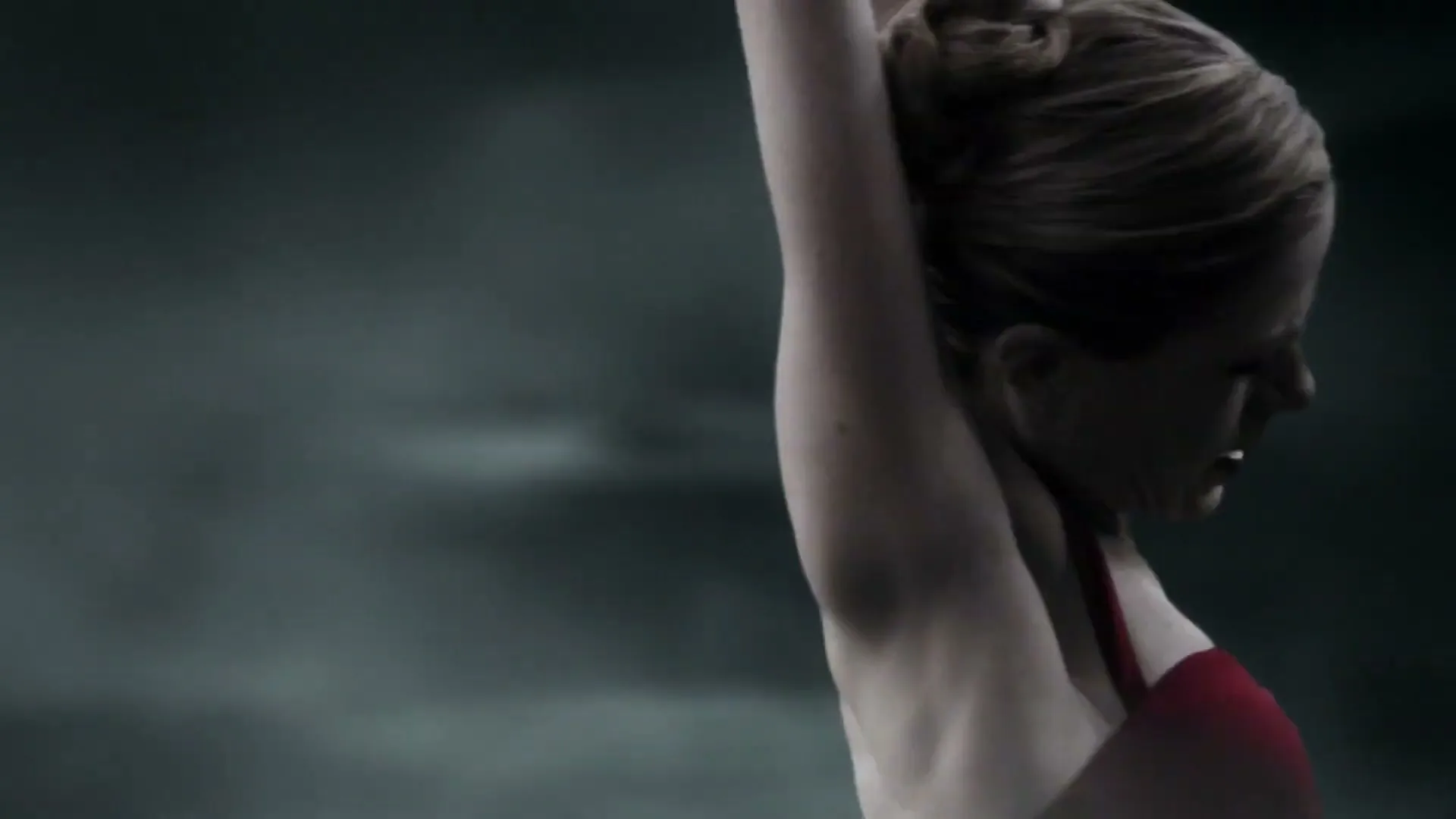 A female ballet dancer in a red dress performs a slow, expressive movement, raising her arms and looking upwards, against a dark, smoky background.