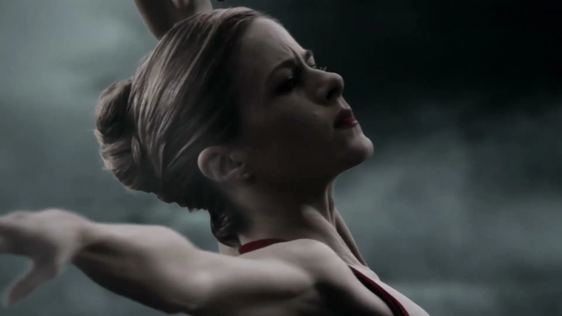 A female ballet dancer with hair in a bun, wearing a red costume, performs a graceful movement against a dark, cloudy background.