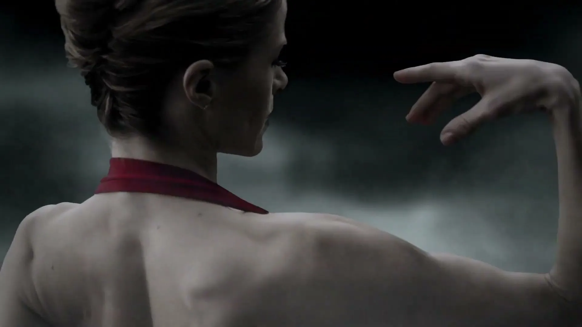 A female ballet dancer in a red leotard performs a graceful movement, turning her head to reveal red lipstick against a dark, smoky background.
