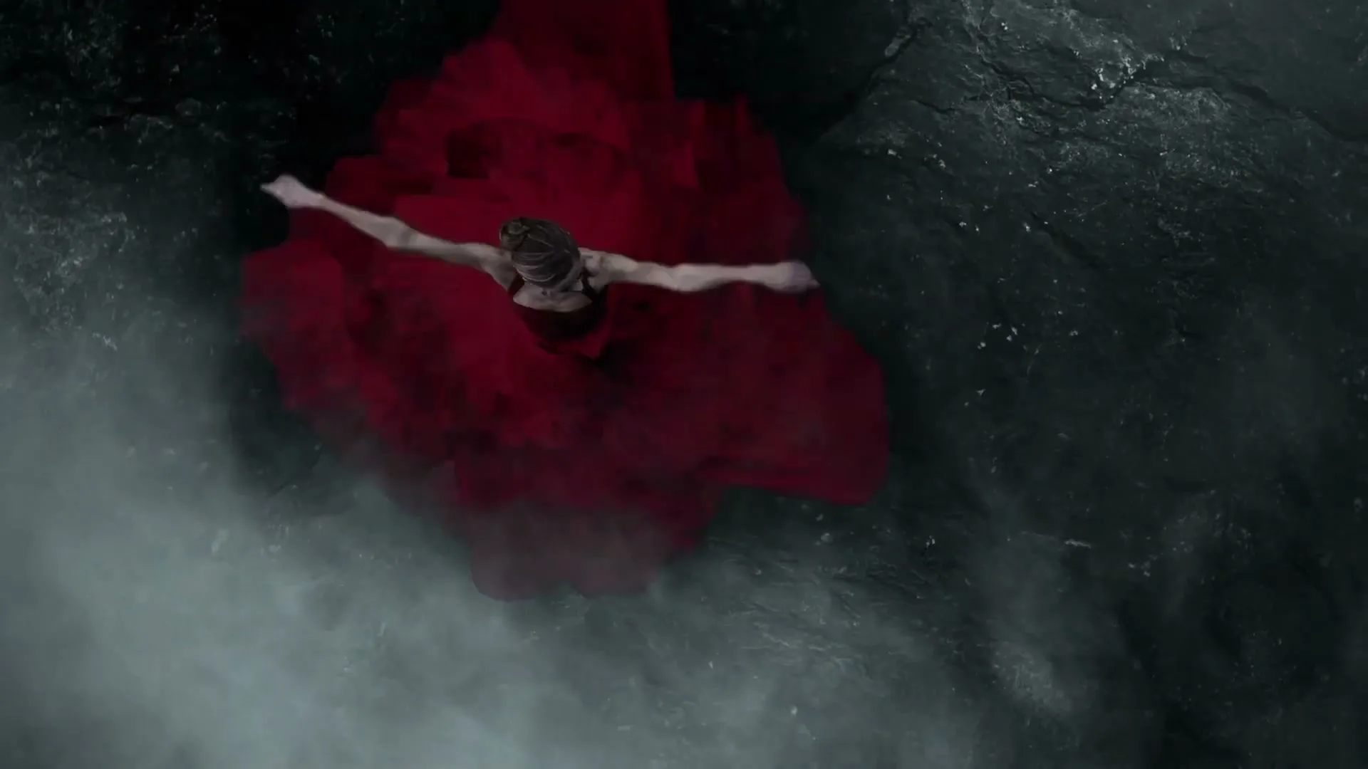 A female ballet dancer in a flowing red dress performs a graceful movement, seen from directly above, against a dark, rocky, misty background.