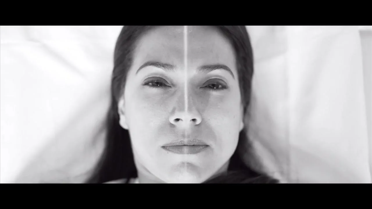 A woman with long dark hair lies on a white bed, eyes closed, then opens them, then the camera pulls back to reveal her in a hospital gown.