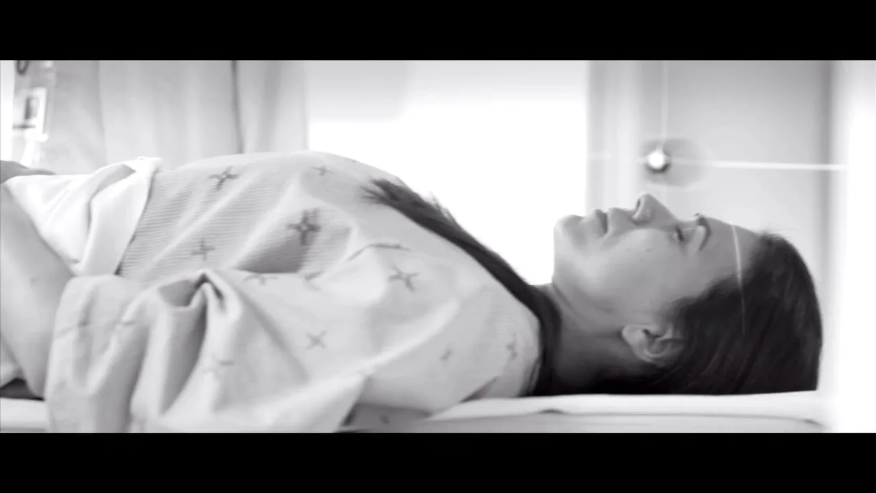 A woman in her 30s, wearing a hospital gown, lies on a bed in a hospital room, looking up with a pained expression.