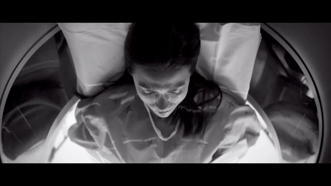 A young woman with dark hair lies on a bed, wearing a hospital gown, as she is slowly moved into an MRI machine in a hospital room.