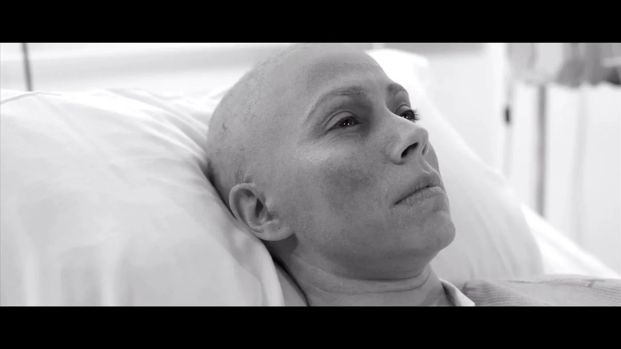 A bald woman with her eyes closed lies in a hospital bed, slowly opening her eyes and looking up, in a black and white shot.
